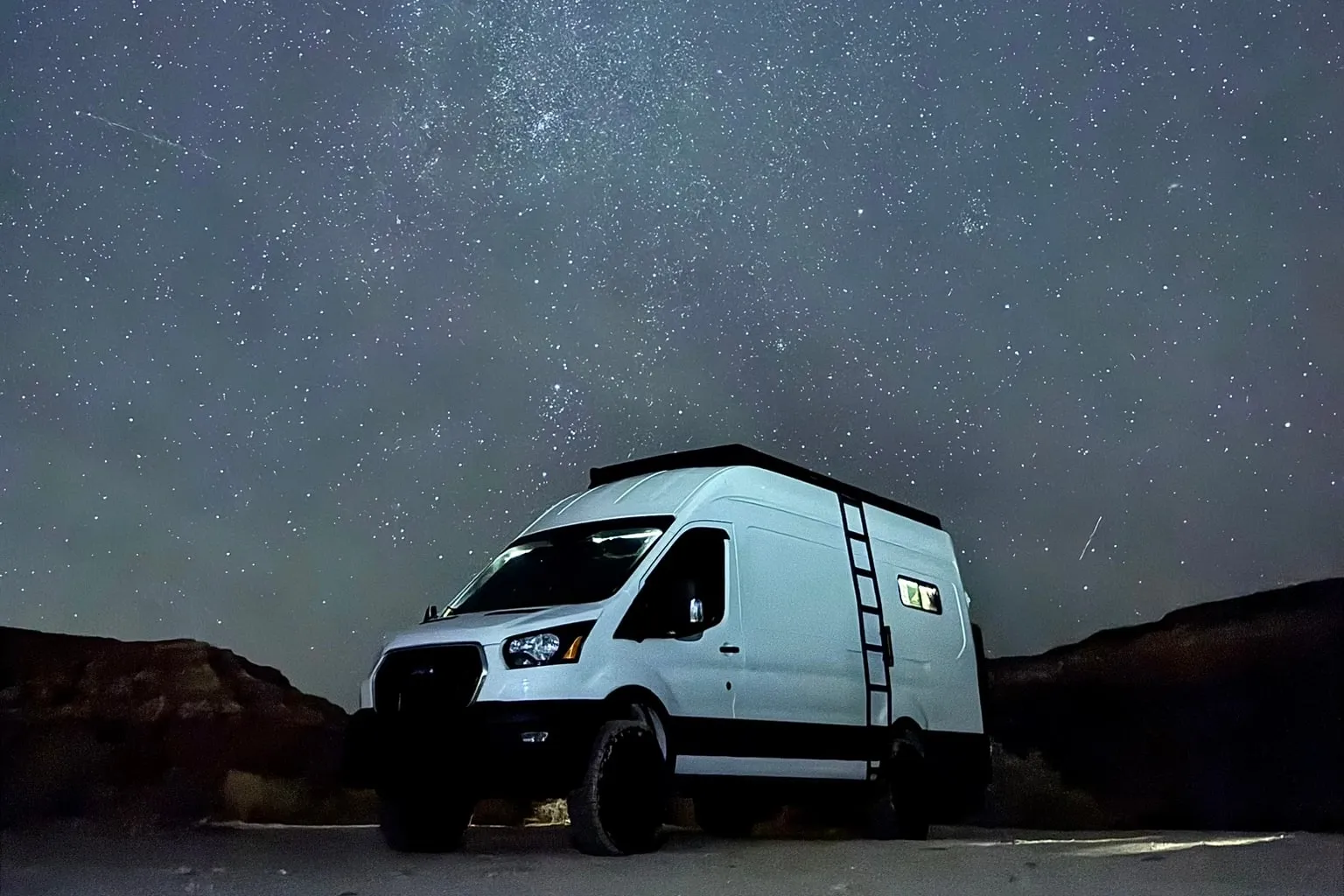 A white Ford Transit camper van parked alone on a desert flat at night, low rock ledges either side of it, under a sky thick with stars and the band of the Milky Way overhead.