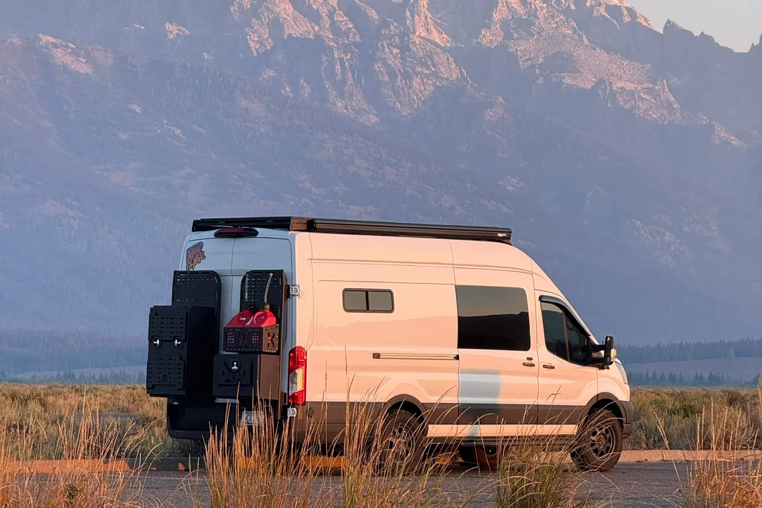 A white Ford Transit camper van parked on a gravel pullout in front of the Teton range at dawn, its rear ladder and gear boxes catching the first orange light, tall dry grass in the foreground.
